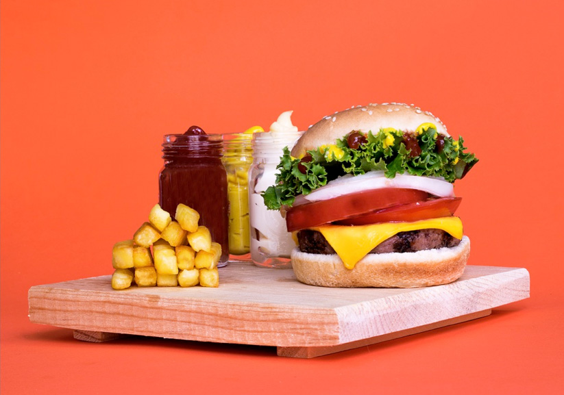 Hamburguesa de carne preparada complementada con lechuga, jitomate y cebolla. Acompañada de papas a la francesa y aderezos de cátsup, mayonesa y mostaza en frasquitos de cristal como decoración